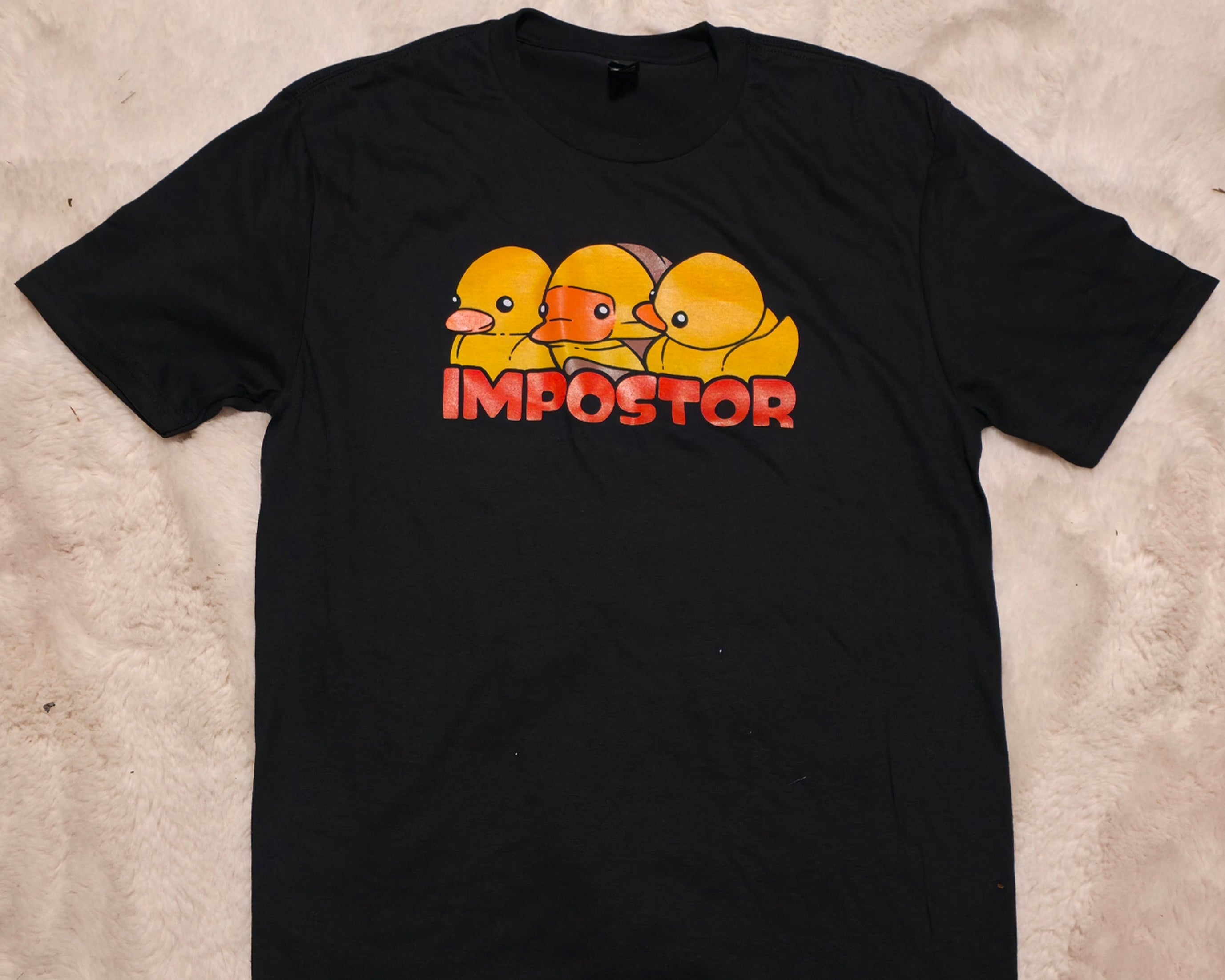 Black t-shirt with yellow rubber duck graphic and 'IMPOSTOR' text on a beige surface